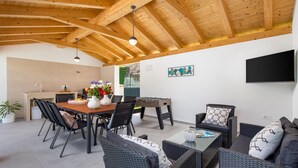 An outdoor dining area with a wooden ceiling, a table with chairs, a sofa, and a television mounted on the wall.