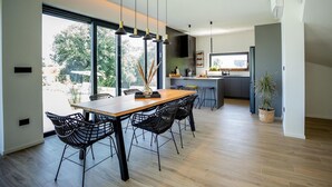Una cucina moderna con una grande isola, un'area pranzo con tavolo in legno e sedie nere, e vista su alberi attraverso ampie finestre.