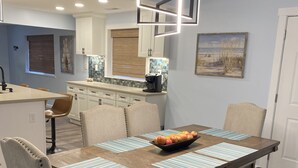A dining area with a wooden table, striped placemats, and a bowl of fruit. There are chairs with cushioned seats and a bench. The room has a modern kitchen in the background with white cabinets and a coffee machine.