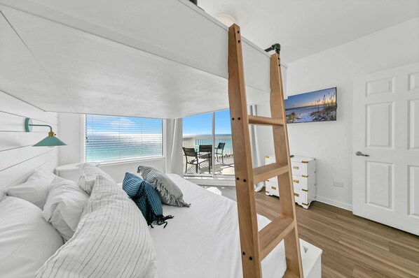 Quarto com cama, escada de madeira, escrivaninha e vista para o oceano atravƩs de grandes janelas.