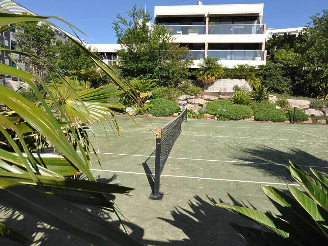 A tennis court with a net in the foreground, surrounded by lush greenery and a modern building in the background.
