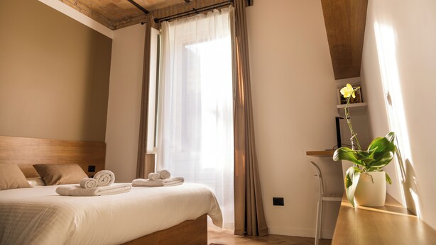 Habitación de hotel con cama, cabecera de madera, escritorio con planta y ventana con cortinas.