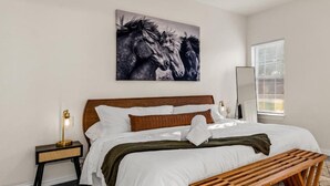 A bedroom with a bed, a bedside table, a floor lamp, a wooden bench, a ceiling fan, a window with blinds, and a wall-mounted artwork of horses.