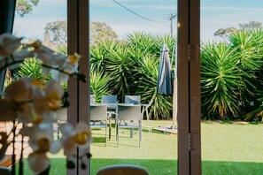 A view through a glass door into a patio area with a table and chairs, surrounded by lush green plants. - The Starfish Cottage - 250m to bay beach (Rye)