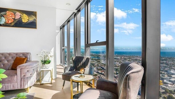 Quarto em um prédio alto com vista para a cidade, uma janela grande, um sofá, uma cadeira e uma mesa pequena.