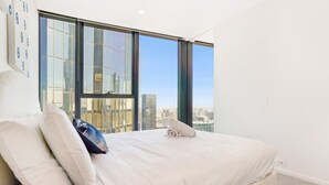 Une chambre d’hôtel avec un grand lit, une vue sur le paysage urbain grâce à de grandes fenêtres du sol au plafond et un mur blanc.