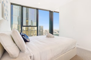 A hotel room with a large bed, white bedding, and a view of a cityscape through floor-to-ceiling windows.