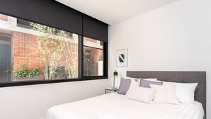 Ein modernes Schlafzimmer mit einem großen Bett, einem Nachttisch, einer Lampe und einem Fenster mit Blick auf ein Ziegelgebäude und Bäume.