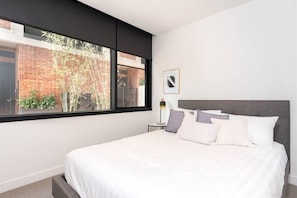 A modern bedroom with a large bed, a nightstand, a lamp, and a window with a view of a brick building and trees.