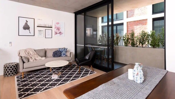 Ein modernes Wohnzimmer mit Sofa, Couchtisch und Essbereich mit Tisch und Stühlen. Auf dem Balkon befinden sich Pflanzen, an den Wänden Kunstwerke.