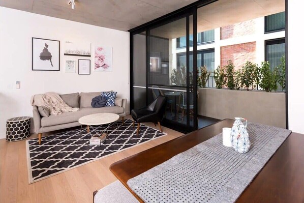 A modern living room with a sofa, coffee table, and a dining area with a table and chairs. There are plants on the balcony and artwork on the walls.