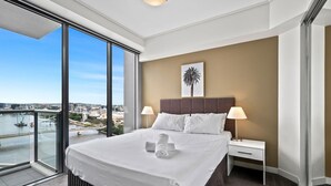 A hotel room with a large bed, a nightstand, a lamp, a view of a cityscape through a balcony, and a framed picture on the wall.