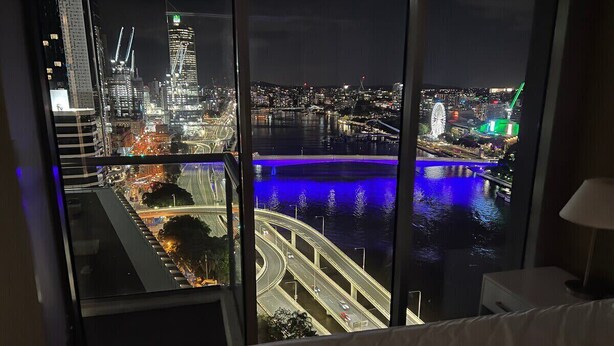 A hotel room with a large window overlooking a cityscape at night, featuring illuminated buildings and a bridge.