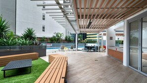 An outdoor patio area with wooden benches, a pool, and a modern building in the background.