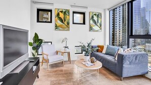 Un soggiorno moderno con una TV a schermo piatto, un divano grigio, una poltrona bianca e un tavolino da caffè. Tre quadri appesi alla parete e un'ampia finestra con vista sulla città.
