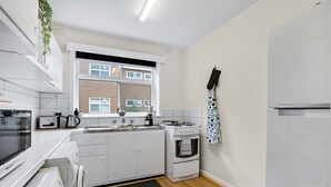 Une cuisine de taille réduite, avec des meubles blancs, un four encastré, une plaque de cuisson et une fenêtre donnant sur un immeuble en briques.