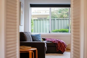 Ein Wohnzimmer mit einer Couch, einem kleinen Tisch und einem Fenster mit Blick auf einen grünen Zaun und Bäume.