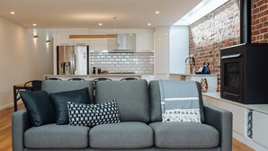 A modern living room with a grey sofa, a kitchen in the background, and a brick wall. - SENSATIONAL Renovated Cottage in Beautiful Seddon (Seddon)