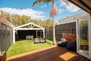 Un patio trasero con terraza de madera, una pérgola blanca, una mesa de comedor y pufs.