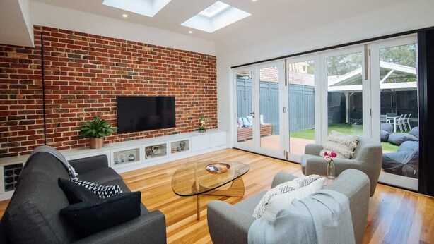 A modern living room with a brick wall, a flat-screen TV, a glass coffee table, and a sliding glass door leading to an outdoor area. - SENSATIONAL Renovated Cottage in Beautiful Seddon (Seddon)