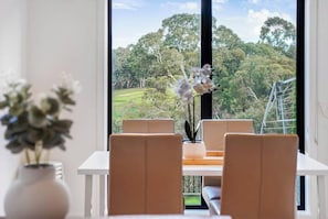 A dining area with a table and chairs, a vase with flowers, and a view of a green landscape through large windows.