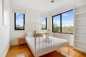 Une chambre avec un lit blanc, une petite table de chevet en bois, une armoire blanche et de grandes fenêtres donnant sur un paysage verdoyant.