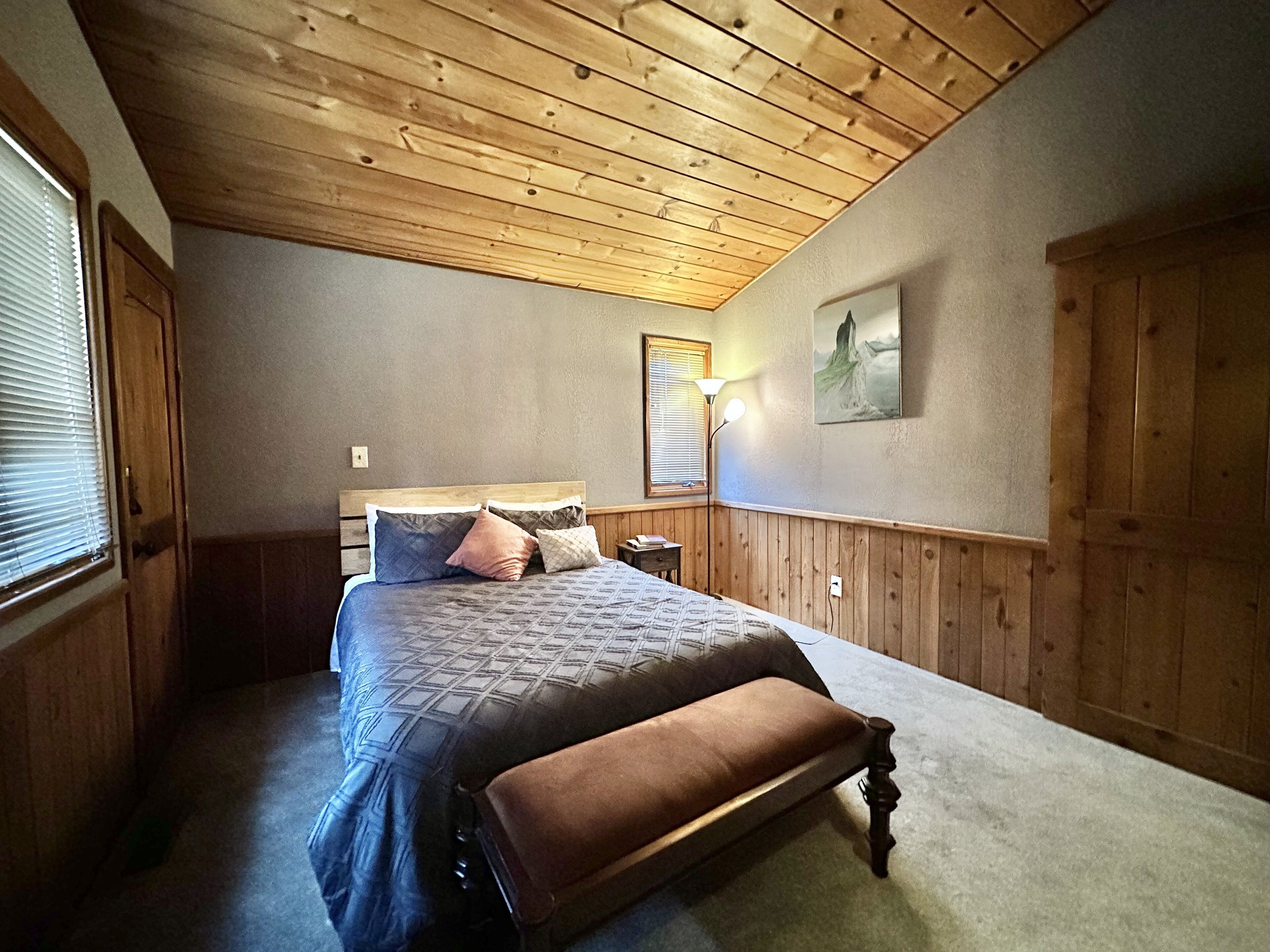 Une chambre avec un plafond en bois, un lit recouvert d’une courtepointe, un banc, une table de chevet, une lampe et un tableau accroché au mur.