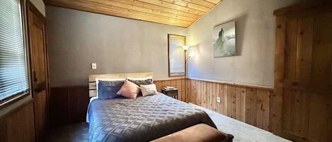Une chambre avec un plafond en bois, un lit recouvert d’une courtepointe, un banc, une table de chevet, une lampe et un tableau accroché au mur.
