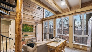 Un interior acogedor con techo de madera, un sofá, una mesa de comedor y ventanas amplias con vistas a árboles.