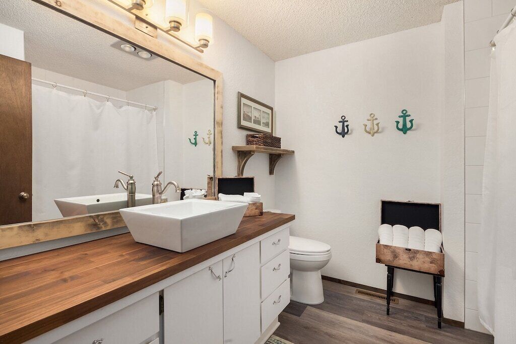 A bathroom with a wooden vanity, a large mirror, a white sink, and a toilet. There are decorative wall anchors and a small shelf with towels.