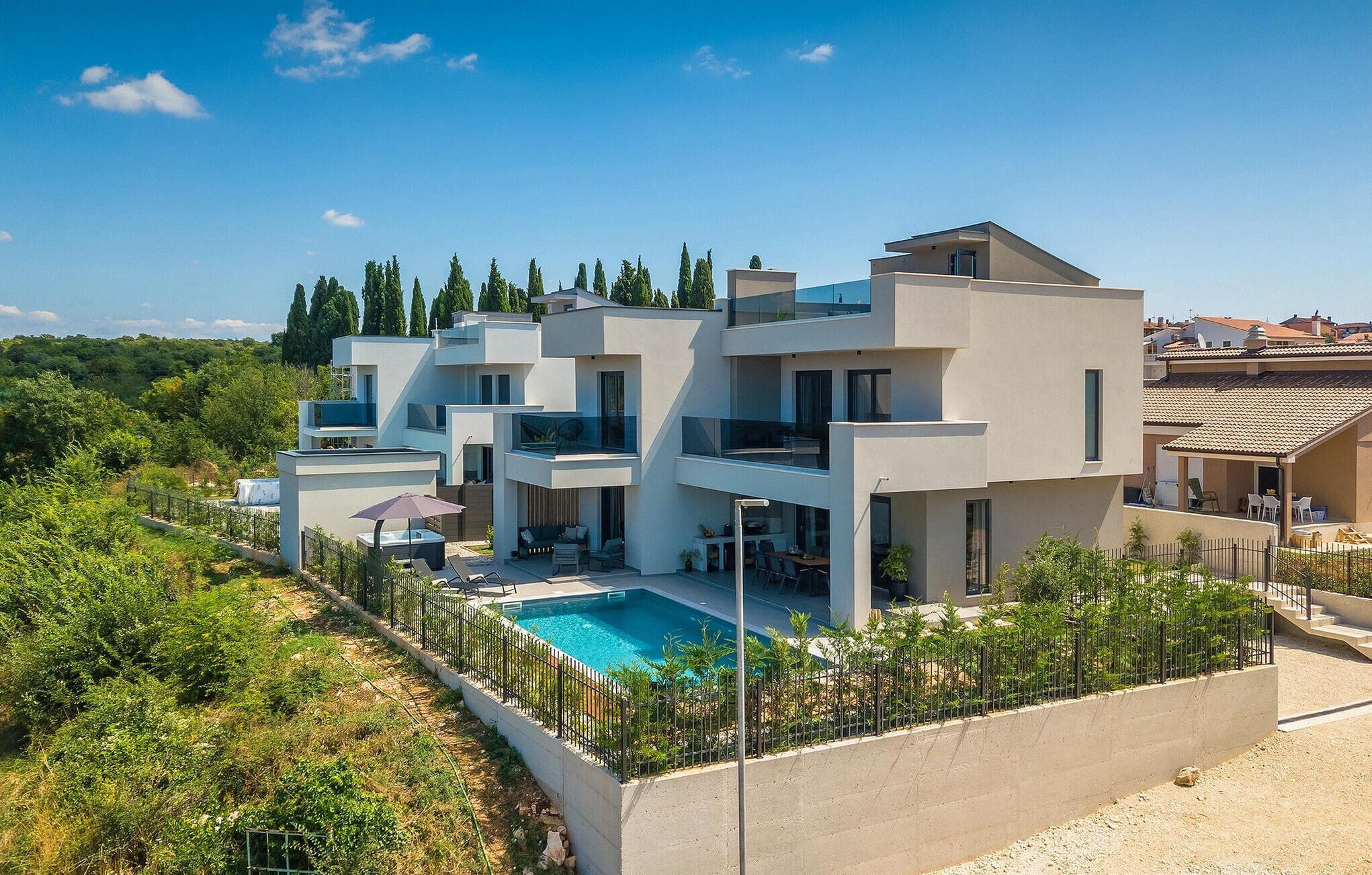 A modern multi-story house with a swimming pool, surrounded by greenery and a clear sky.