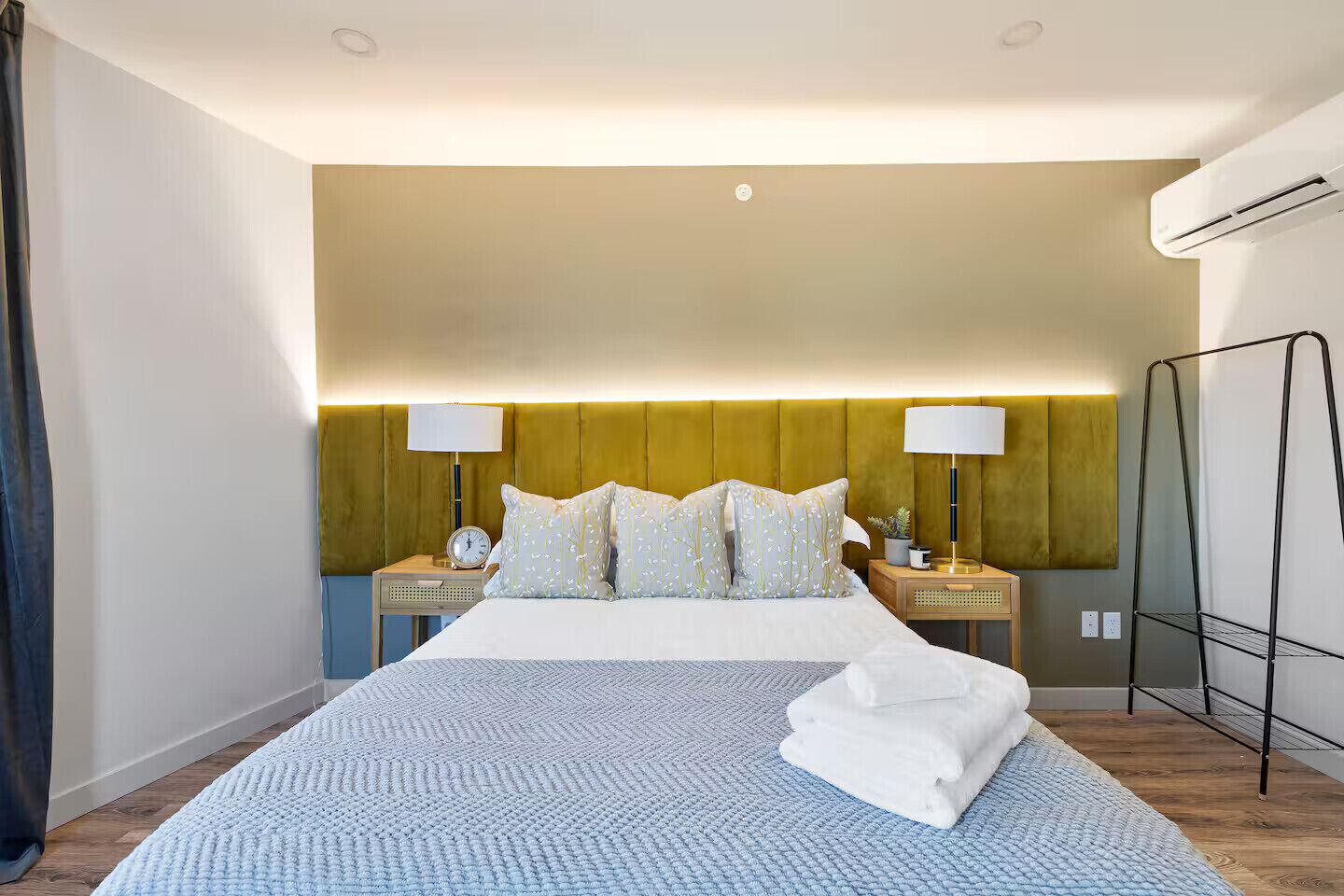 A hotel room with a bed, bedside tables, lamps, an air conditioning unit, and a wall-mounted shelf.