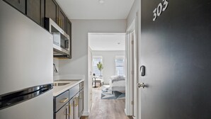 A modern kitchen with a built-in microwave, a stainless steel refrigerator, and a wooden floor leading to a bedroom with a bed and a window. - Cottage Suites (Middletown)