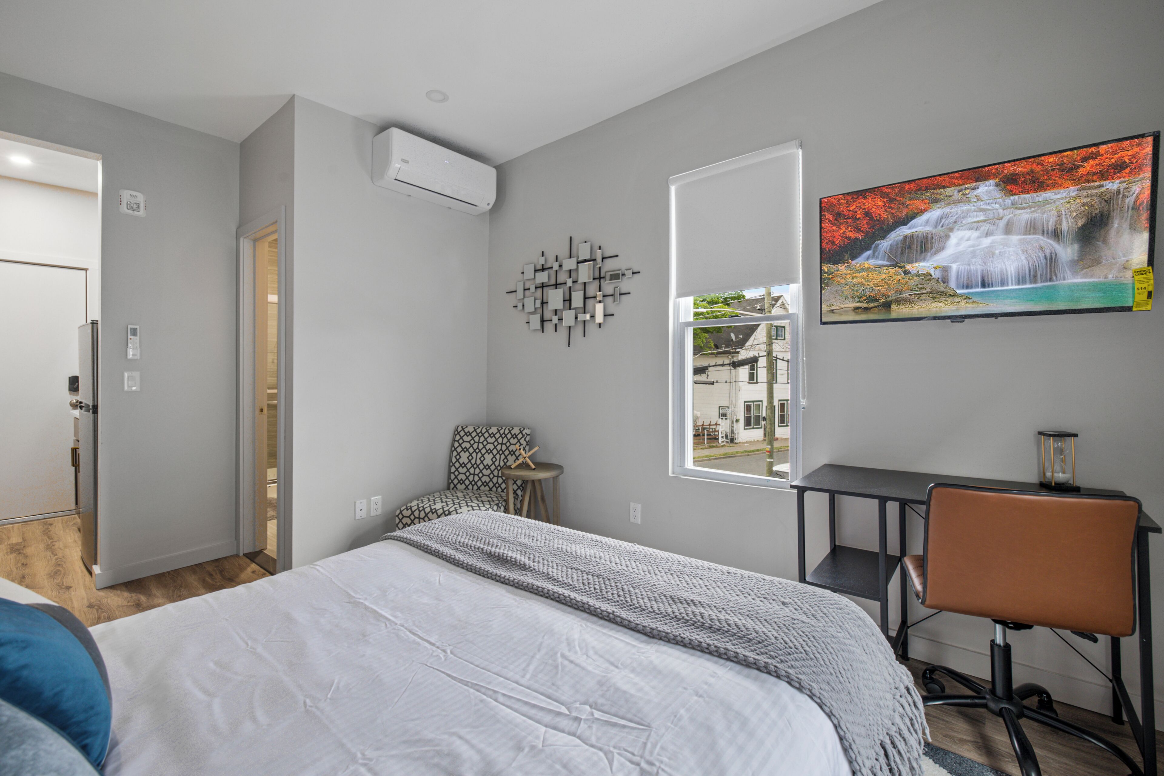 Un dormitorio con cama, escritorio, silla, ventana y televisor instalado en la pared.