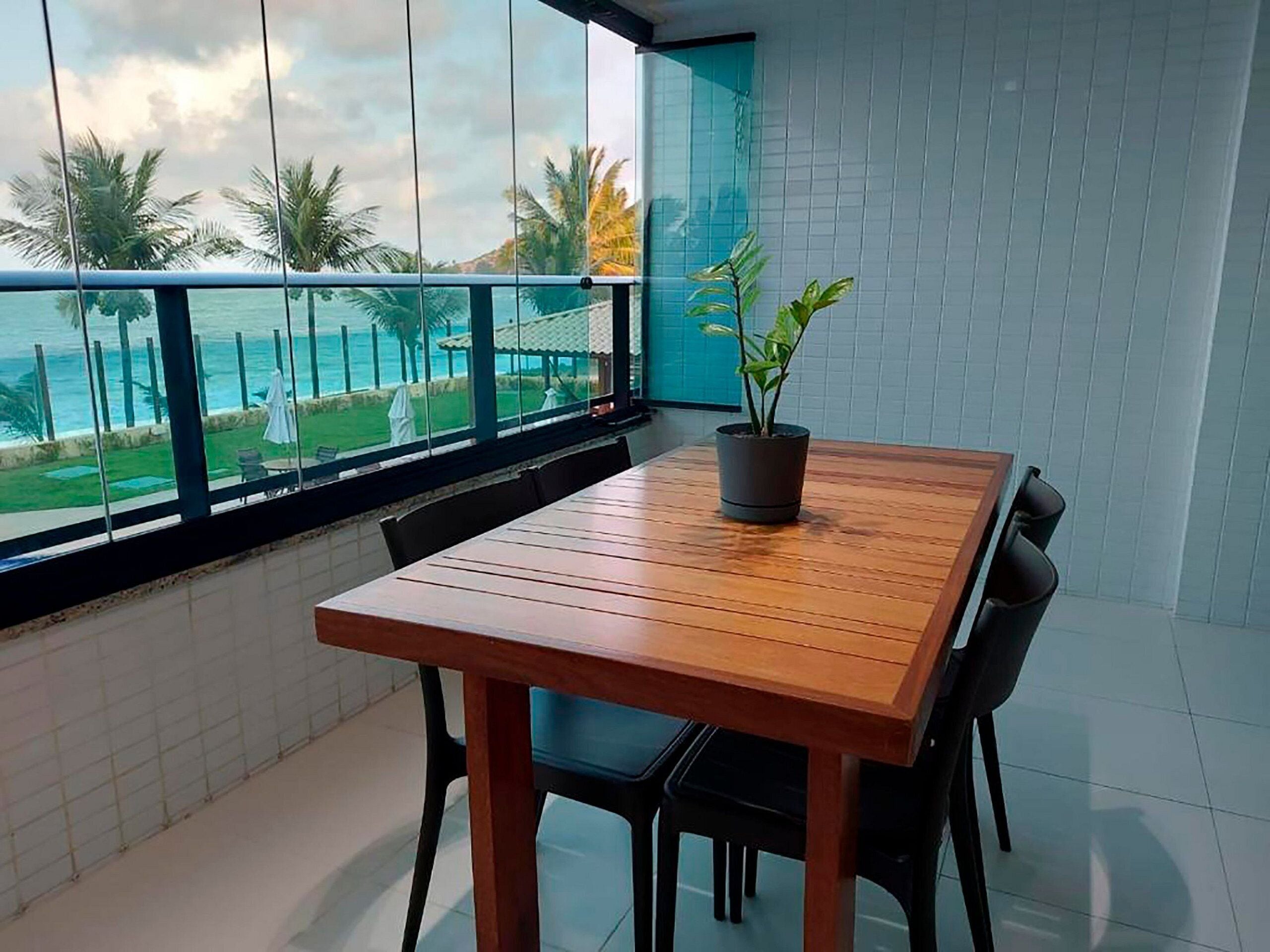 A balcony with a wooden table and chairs, a potted plant, and a view of the ocean and palm trees.