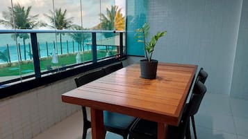 A balcony with a wooden table and chairs, a potted plant, and a view of the ocean and palm trees.