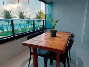 Een balkon met een houten tafel en stoelen, een potplant en uitzicht op de oceaan en palmbomen.