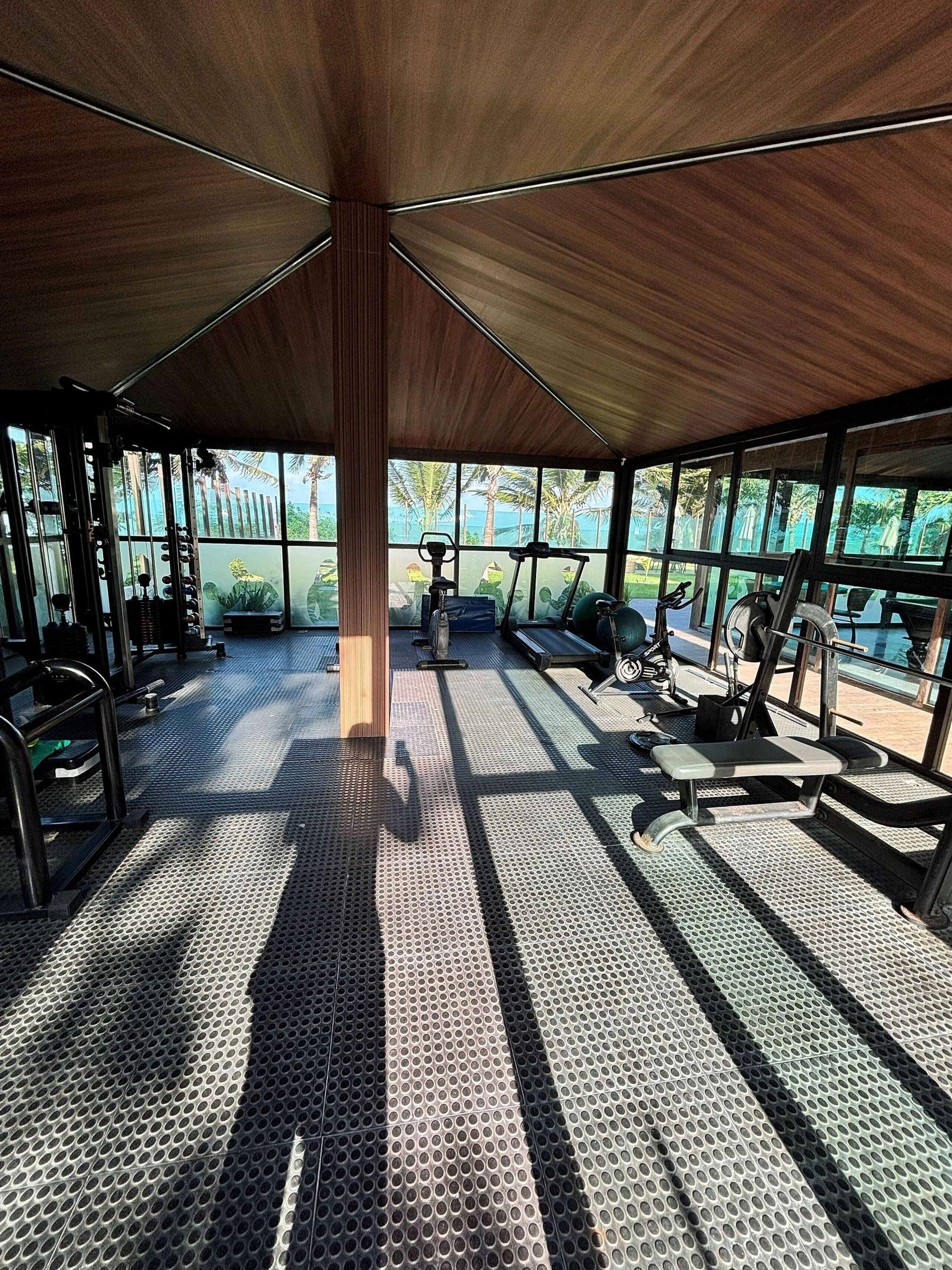 Un gimnasio techado con equipo de ejercicio, suelo estampado y ventanas amplias que ofrecen vista a palmeras y al mar.