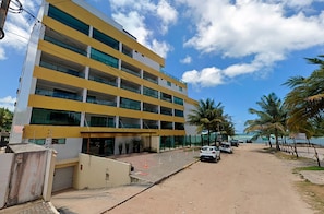 Un immeuble à plusieurs étages, doté d’une façade jaune et de grandes fenêtres, situé près d’une plage bordée de palmiers, sous un ciel dégagé.