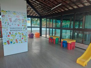 A children's play area with colorful tables and a yellow slide, under a large glass window. - Flat Frente Mar - Praia de Tabatinga (Conde)