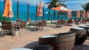 Un salon de plage avec des fauteuils et des tables en osier, des parasols orange et une vue sur l’océan.
