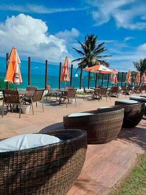 Un salon de plage avec des fauteuils et des tables en osier, des parasols orange et une vue sur l’océan.