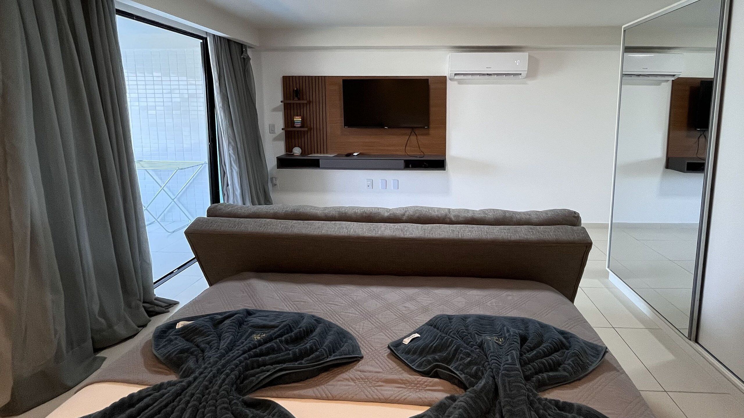 A modern hotel room with a large bed, a flat-screen TV mounted on the wall, and a large mirror on one side.