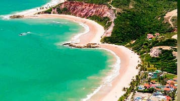 An aerial view of a coastal area with a curved beach, a cliff, and a resort with multiple buildings and a swimming pool.