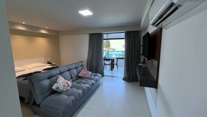Une chambre d’hôtel moderne avec un canapé gris, une petite table, un téléviseur fixé au mur et un balcon offrant une vue sur la mer.