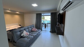 Une chambre d’hôtel moderne avec un canapé gris, une petite table, un téléviseur fixé au mur et un balcon offrant une vue sur la mer.
