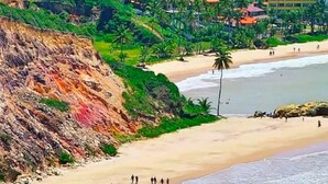 A coastal area with a sandy beach, a rocky cliff, and a building with a red roof in the background. - Flat Frente Mar - Praia de Tabatinga (Conde)