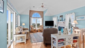 A spacious living room with a large window, a ceiling fan, a flat-screen TV, and a white coffee table. - 5591- Beach, Please! - Soundside Escape in Nags Head | Dog Friendly (Nags Head)