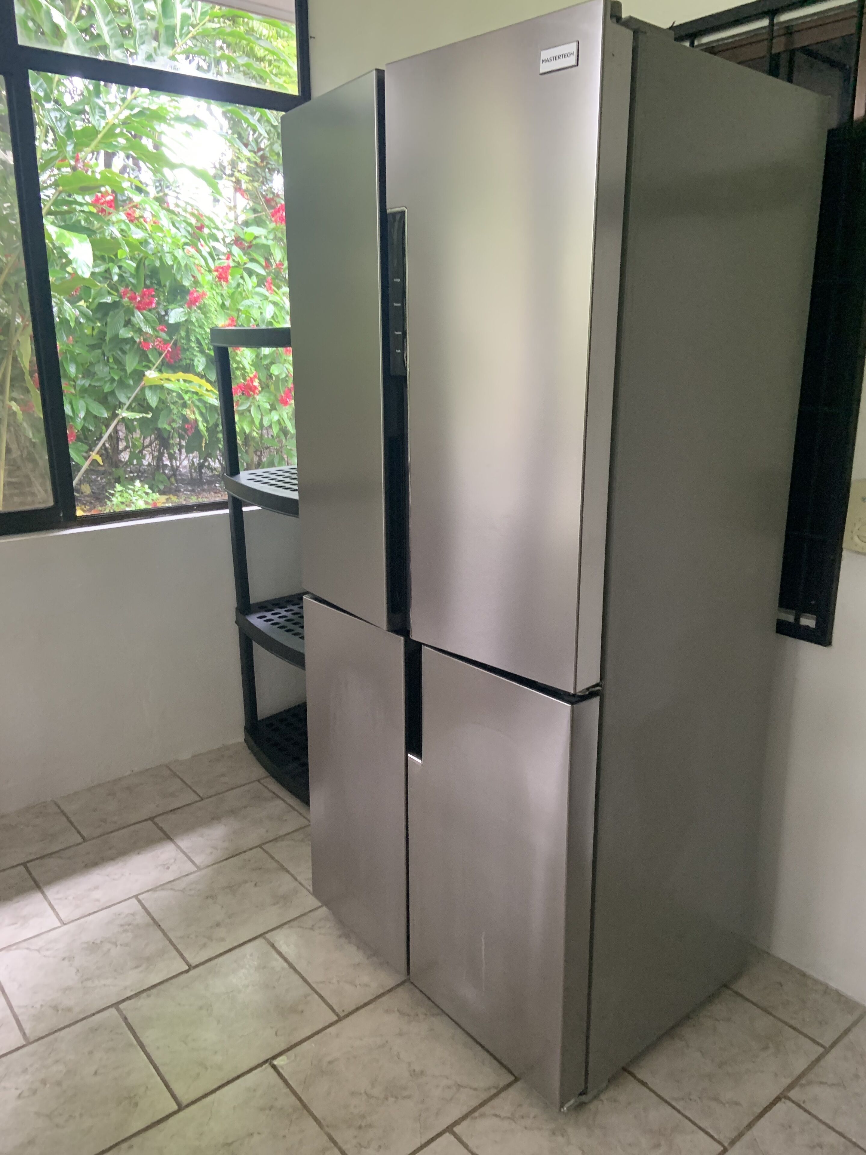 A stainless steel refrigerator in a kitchen with tiled flooring and a window overlooking a garden with red flowers.
