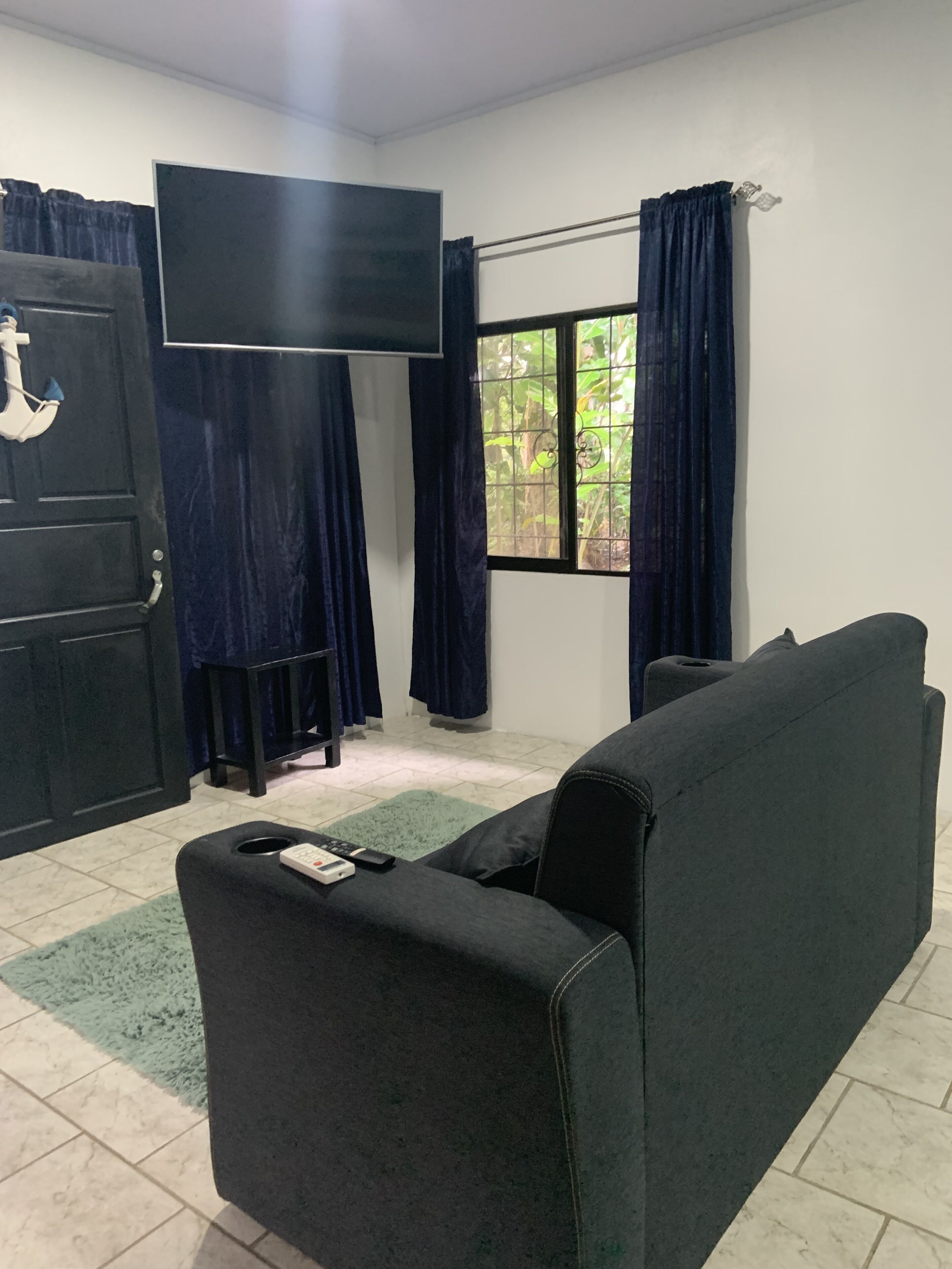 Un salon avec un canapé noir, un téléviseur fixé au mur, une fenêtre avec des rideaux et une porte également dotée d’un rideau.